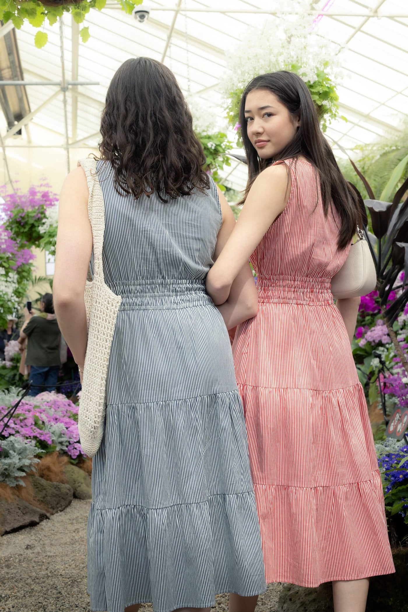 Two models in striped tiered midi dresses — blue and pink — standing together in a flower-filled greenhouse