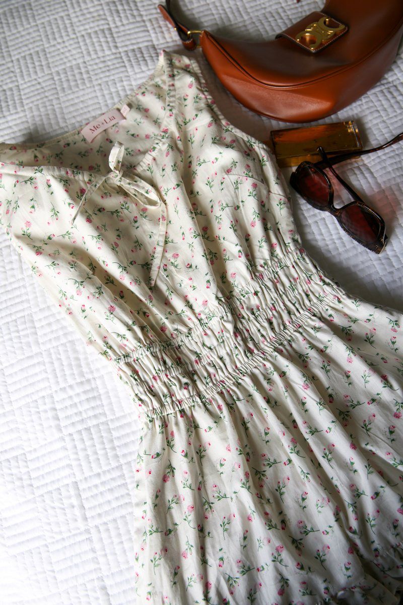 A folded floral cotton dress with a leather bag and sunglasses on a quilted bed