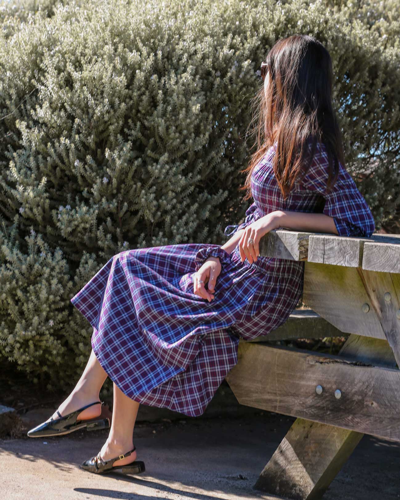 Model seated on a wooden bench in a burgundy plaid midi dress, coastal shrubs in the background